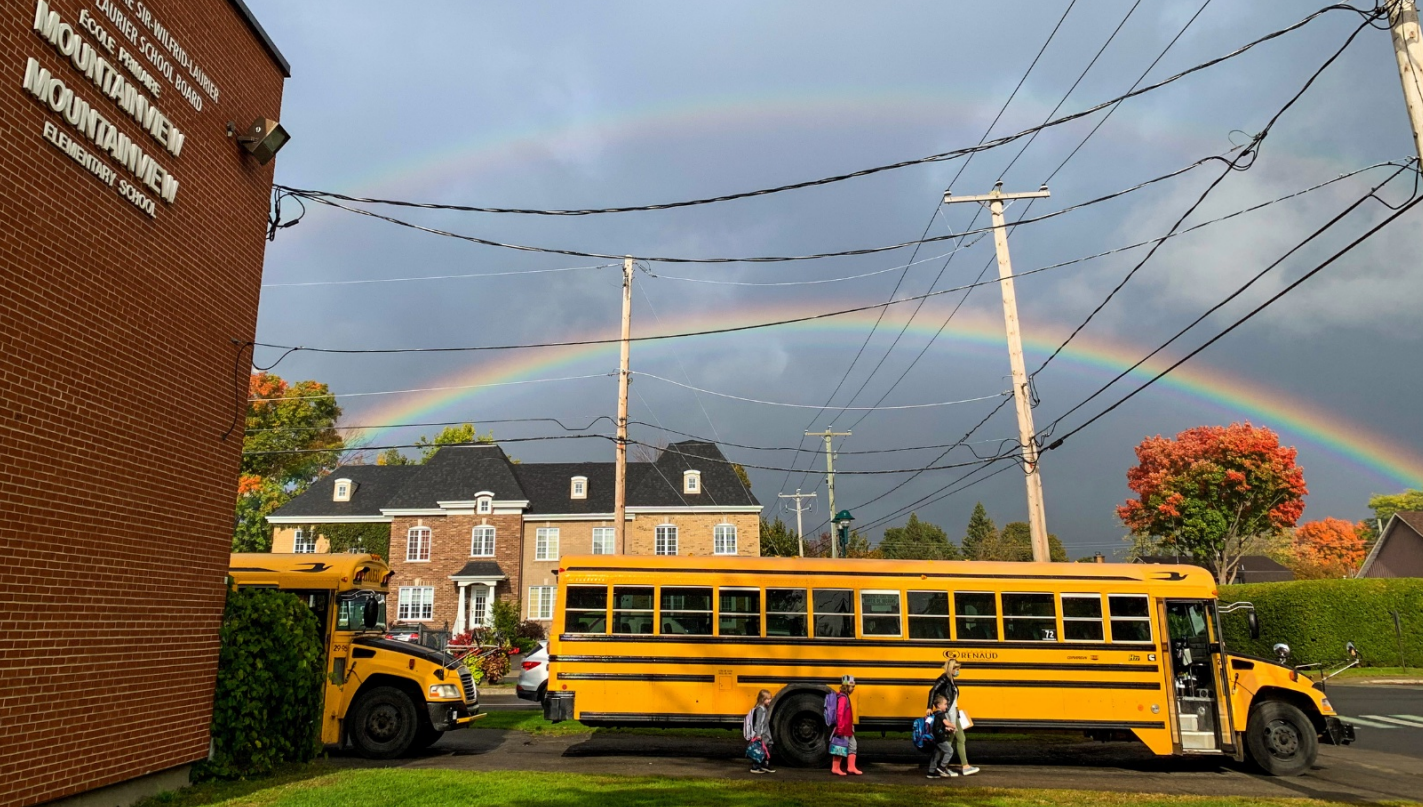 A school bus. A teacher walking with students with a rainbow in the sky over the bus. The words “Our School” are across the image.